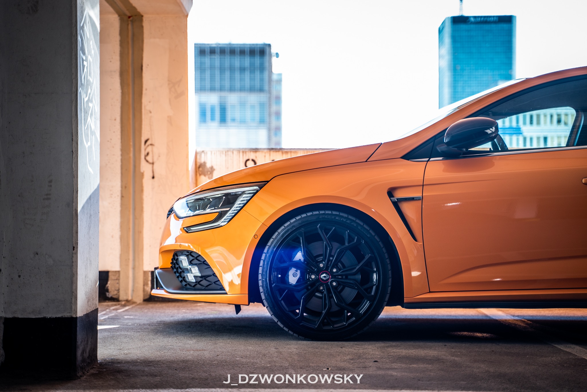 Pomarańczowy samochód Renault Megane RS z czarnymi felgami na tle betonowego parkingu i wieżowców