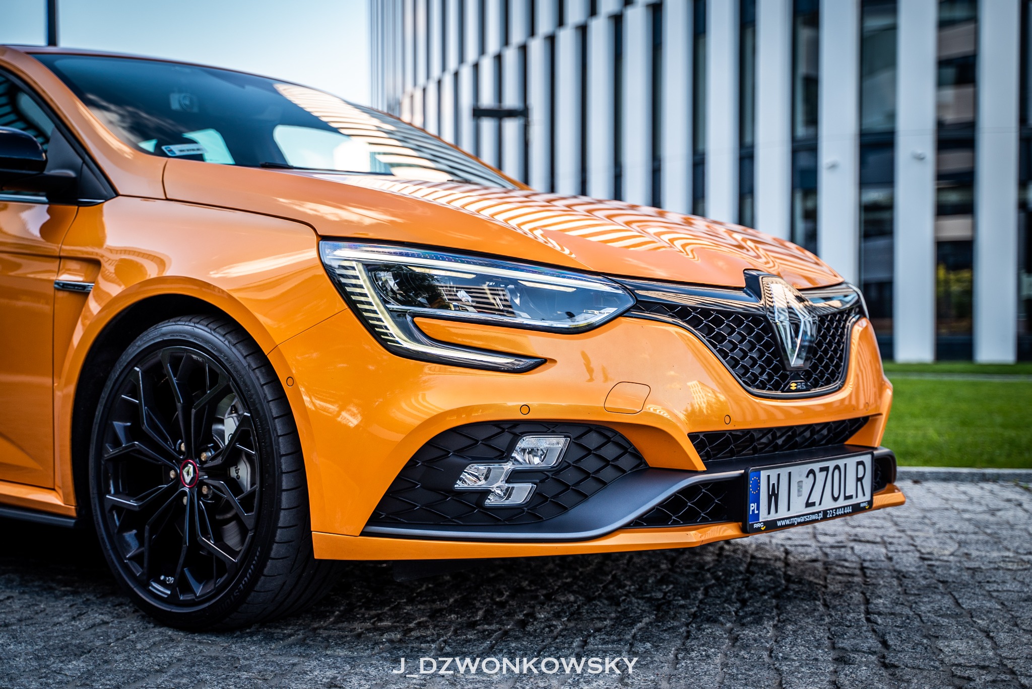 Pomarańczowy samochód marki Renault Megane RS z czarnymi felgami na tle nowoczesnego budynku z białymi pionowymi panelami.