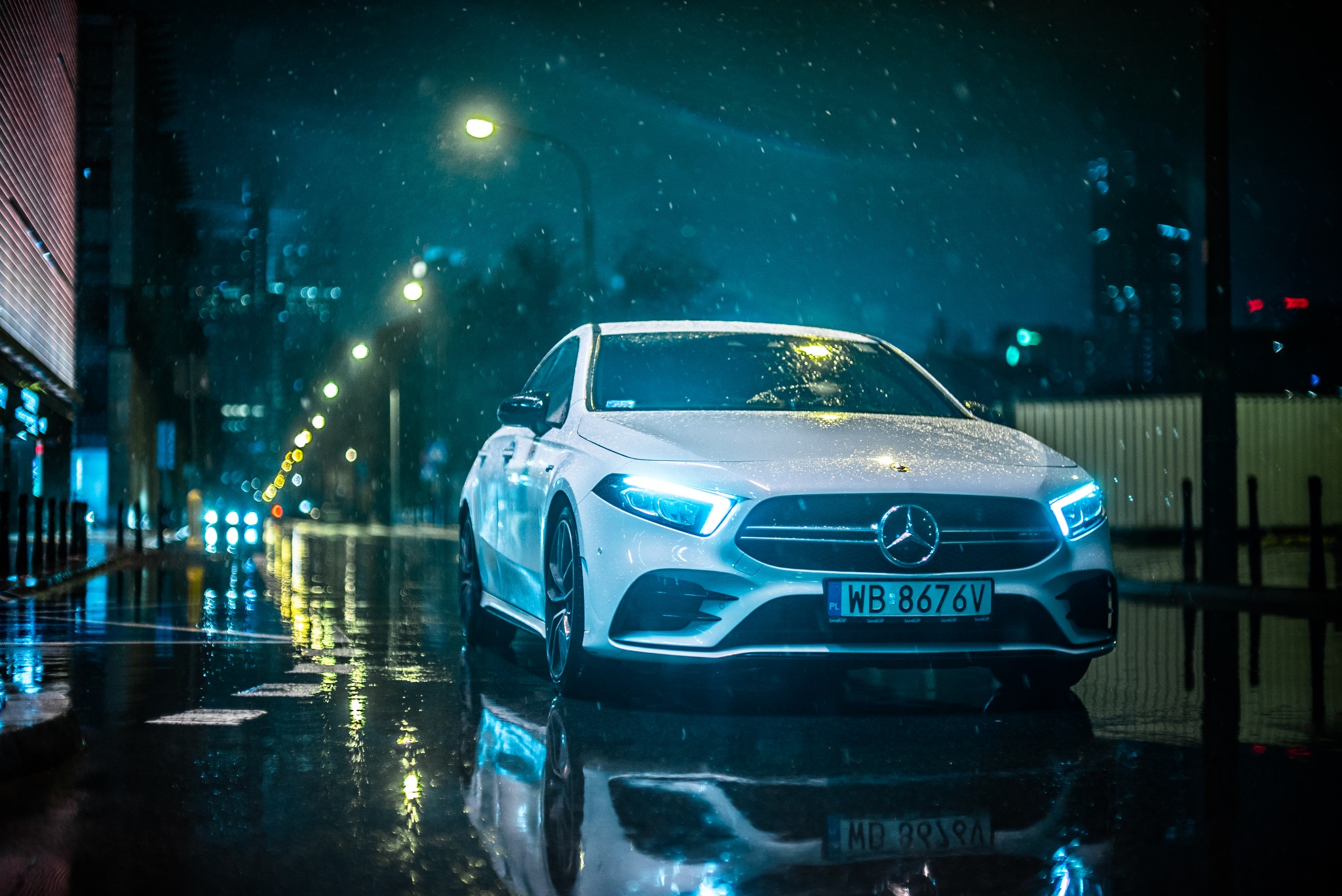 Biały samochód marki Mercedes-Benz A-klasa zaparkowany na mokrej ulicy w nocy, z widocznymi odbiciami świateł miasta i padającego deszczu.