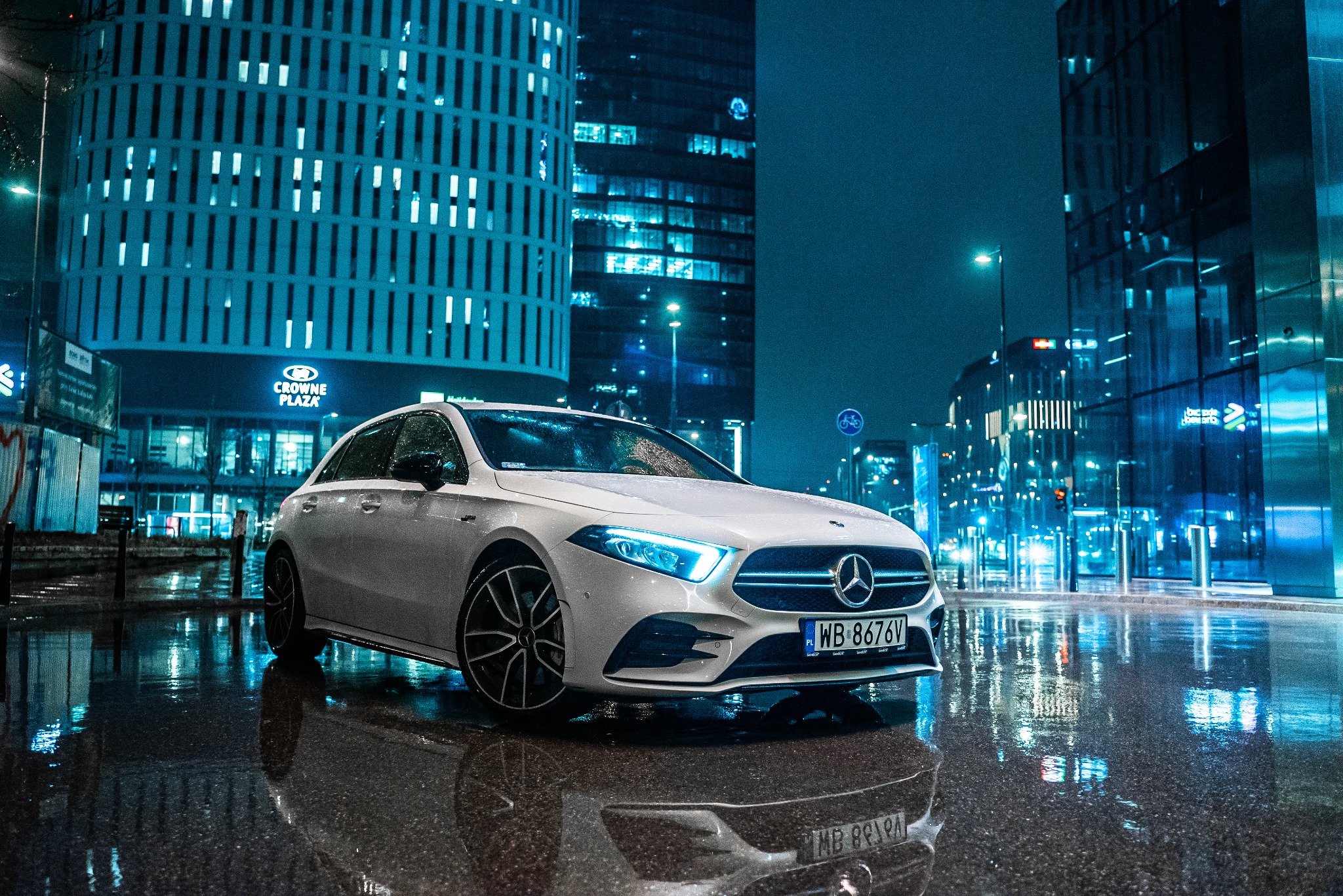 Biały Mercedes A-klasa z włączonymi światłami LED, zaparkowany na mokrej nawierzchni z odbiciami miejskich świateł i budynków w tle, nocna sceneria.