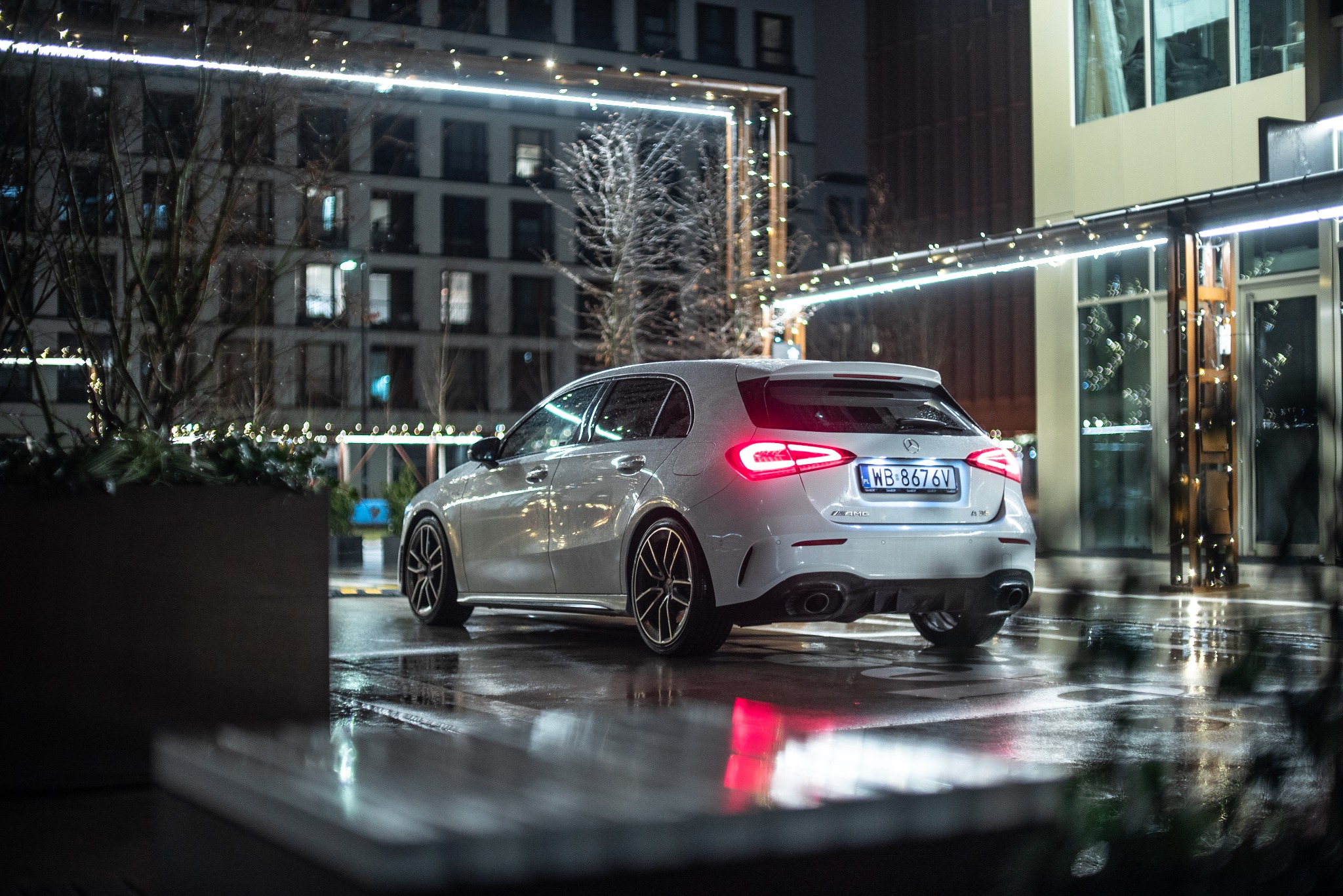 Srebrny samochód marki Mercedes-Benz klasy A, widziany od tyłu na mokrym asfalcie w nocy, z widocznymi światłami tylnymi i tablicą rejestracyjną, na tle oświetlonego budynku.