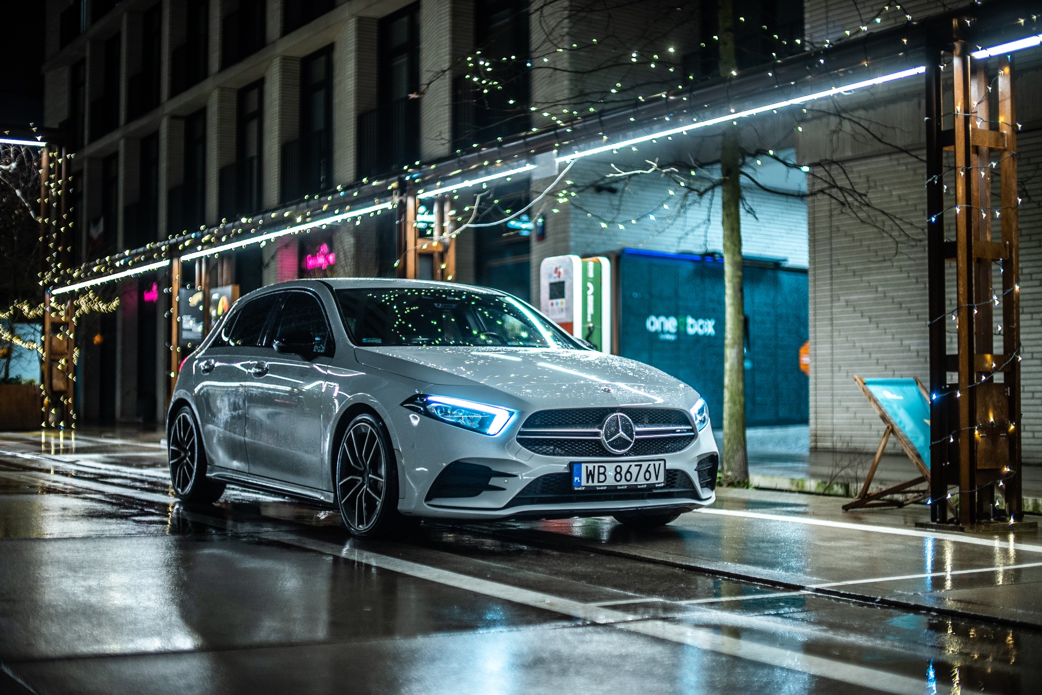 Biały samochód marki Mercedes-Benz klasy A zaparkowany nocą na mokrej ulicy, z odbiciami świateł w kałużach, na tle oświetlonego budynku.