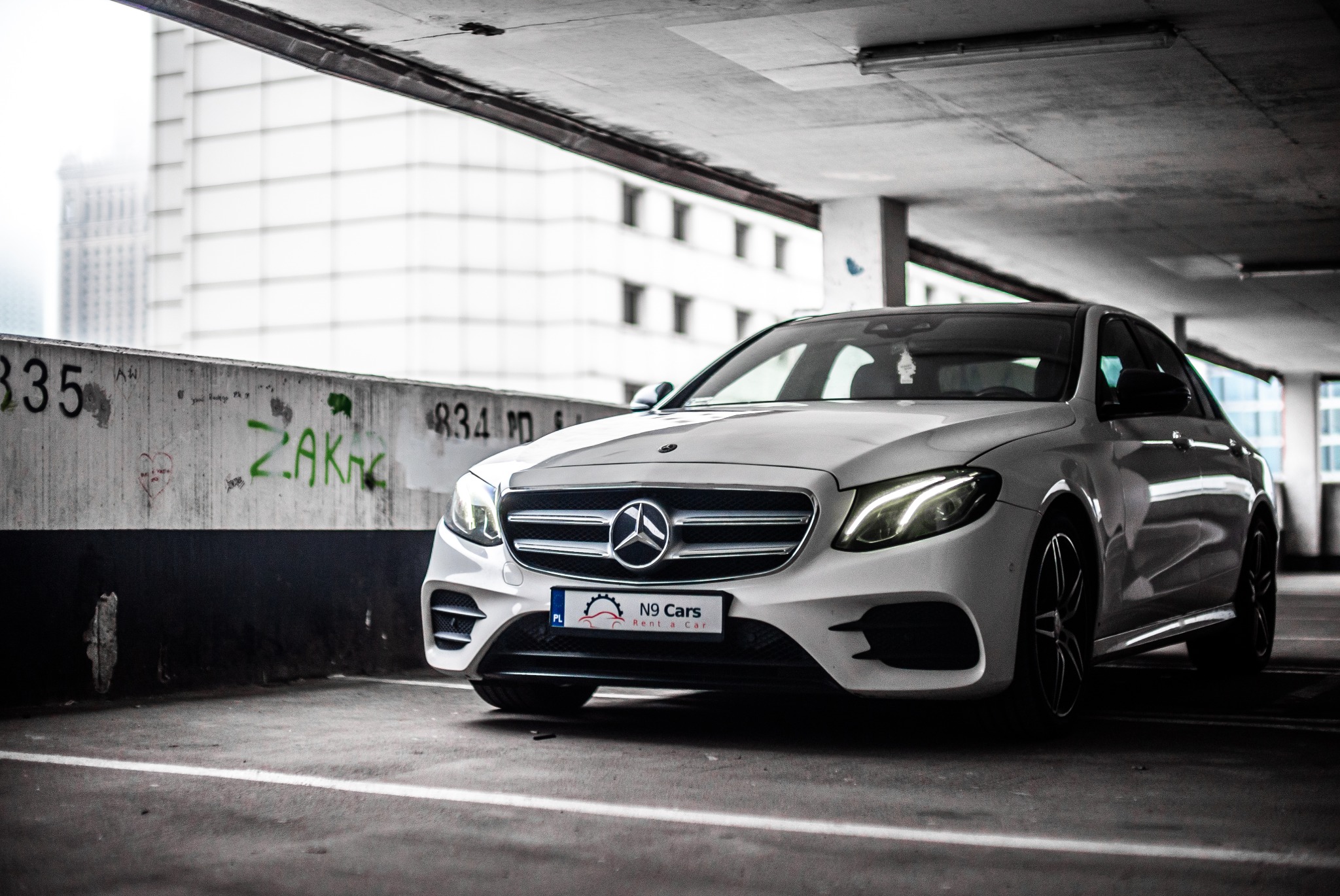 Biały Mercedes E-klasa na parkingu podziemnym, widoczny napis 'N9 Cars Rent a Car' na tablicy rejestracyjnej, w tle budynek biurowy.