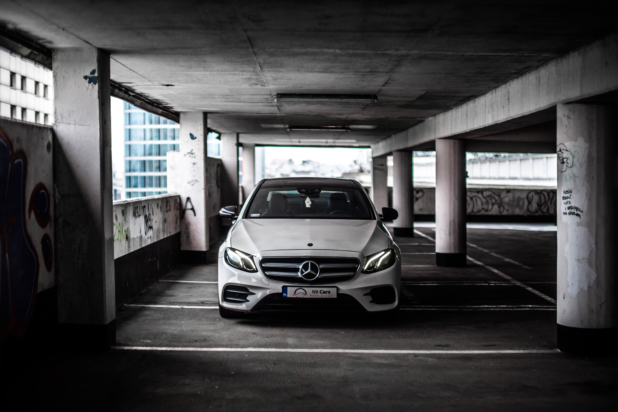 Biały samochód marki Mercedes zaparkowany w garażu podziemnym z graffiti na ścianach i kolumnach, widok z przodu.