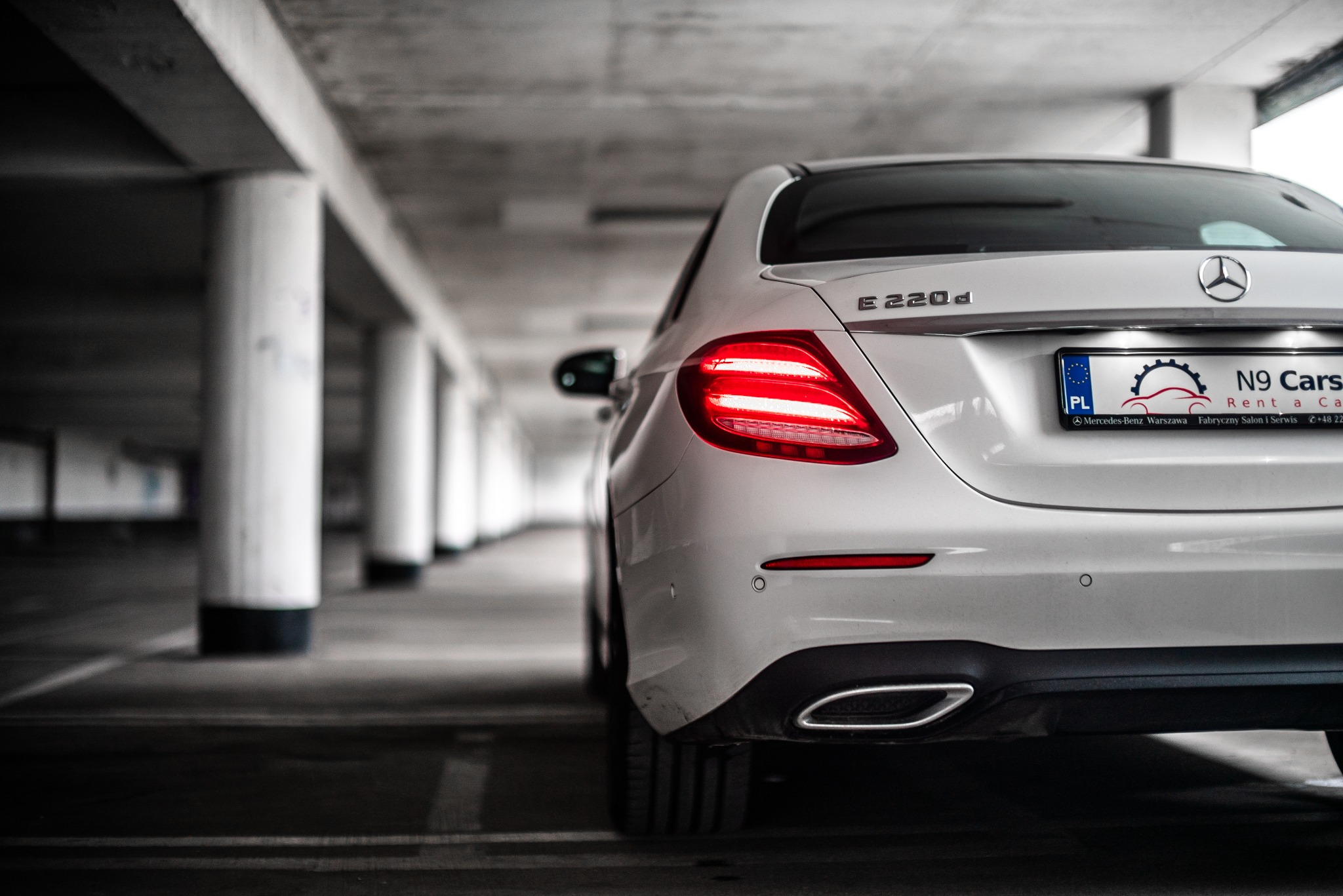 Tył białego Mercedesa E220d z włączonymi światłami, zaparkowanego w garażu podziemnym z rzędem kolumn, widoczna tablica rejestracyjna firmy wynajmującej samochody.