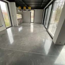 Szara, błyszcząca posadzka w nowym pomieszczeniu z dużymi oknami, widoczne elementy instalacji pod sufitem i na ścianach.