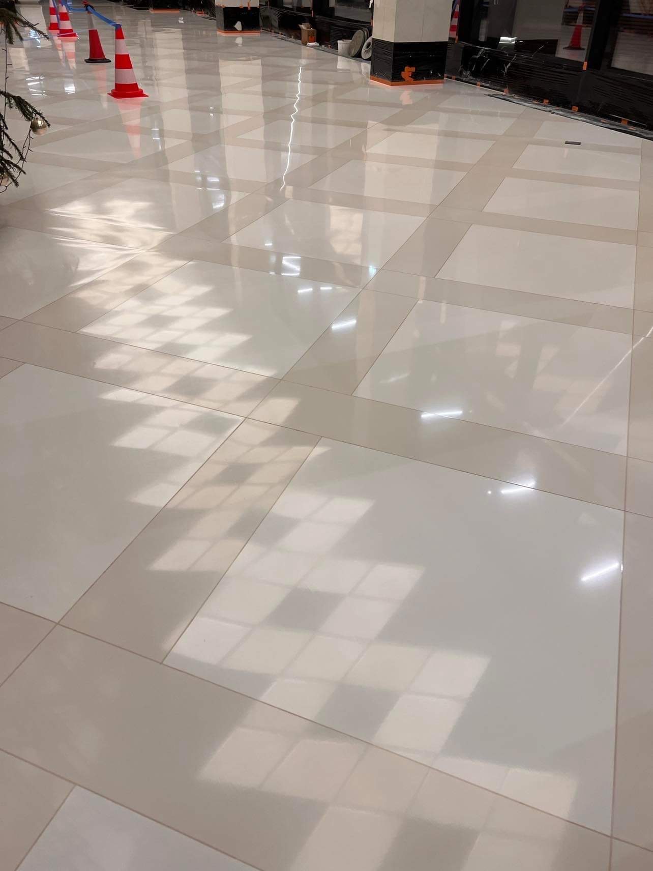 Błyszcząca, jasna posadzka przemysłowa z żywicy epoksydowej, zabezpieczona stożkami ostrzegawczymi podczas schnięcia, widoczne refleksy światła i cień okien.