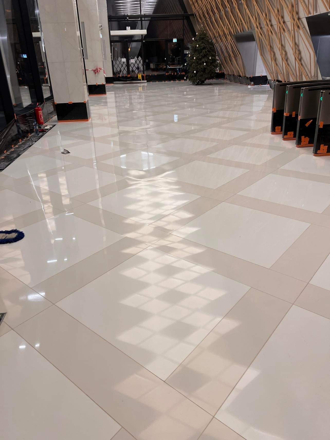 Błyszcząca, jasna posadzka w hali biurowej, ułożona z kwadratowych płyt w odcieniach beżu i bieli, z widocznymi refleksami światła i elementami wyposażenia hali w tle.