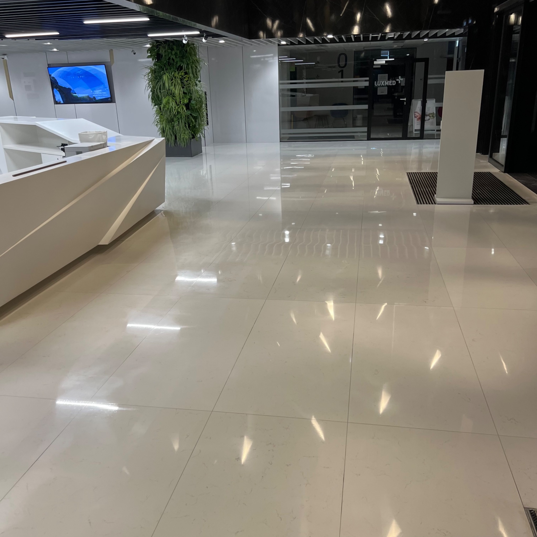 Błyszcząca, jasna posadzka w nowoczesnym biurze Luxmed, z widocznym odbiciem światła i elementami wystroju, takimi jak recepcja i pionowa ściana zieleni.