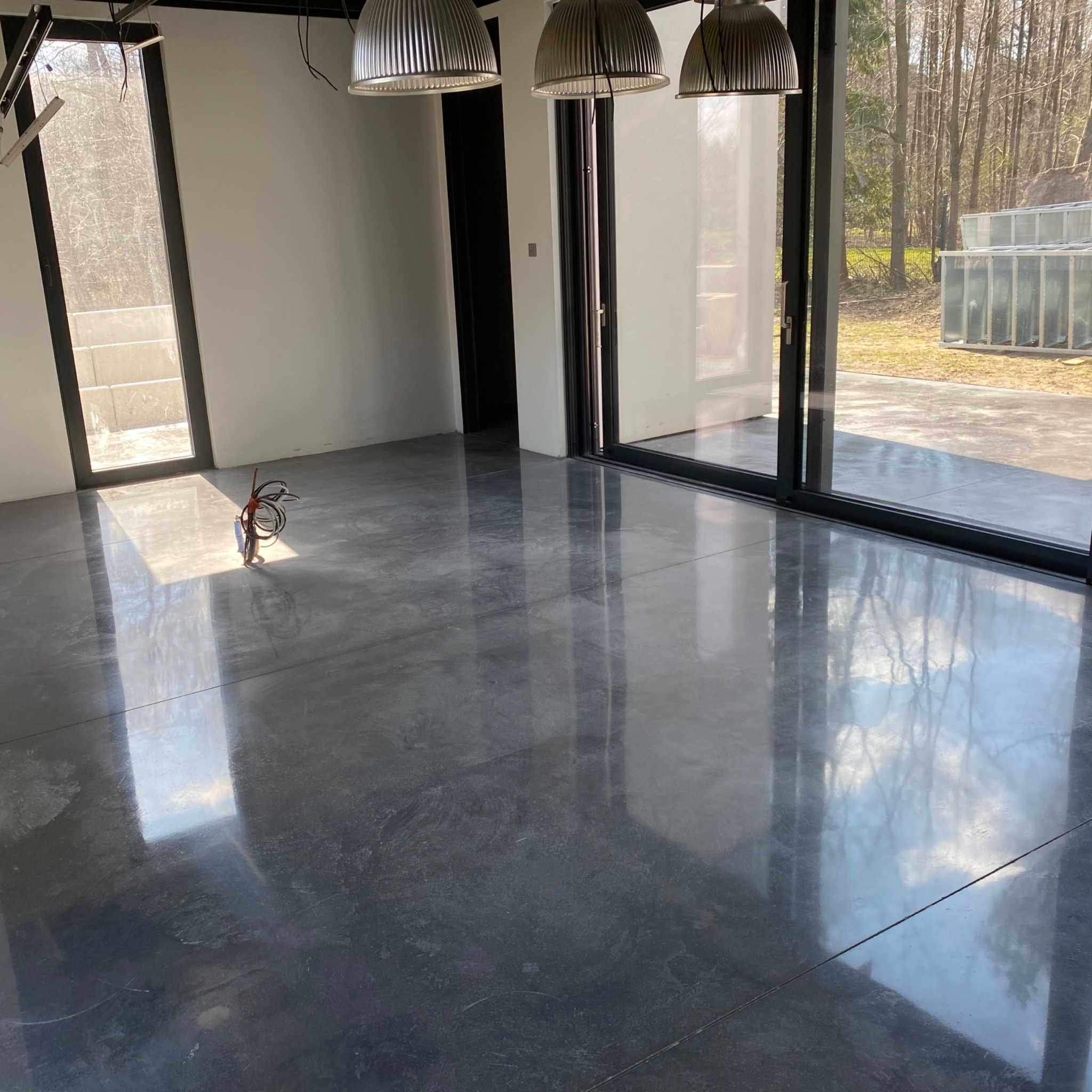 Błyszcząca, szara posadzka w jasnym pomieszczeniu z dużymi oknami, widoczne wiszące lampy i kable instalacyjne przy ścianie.