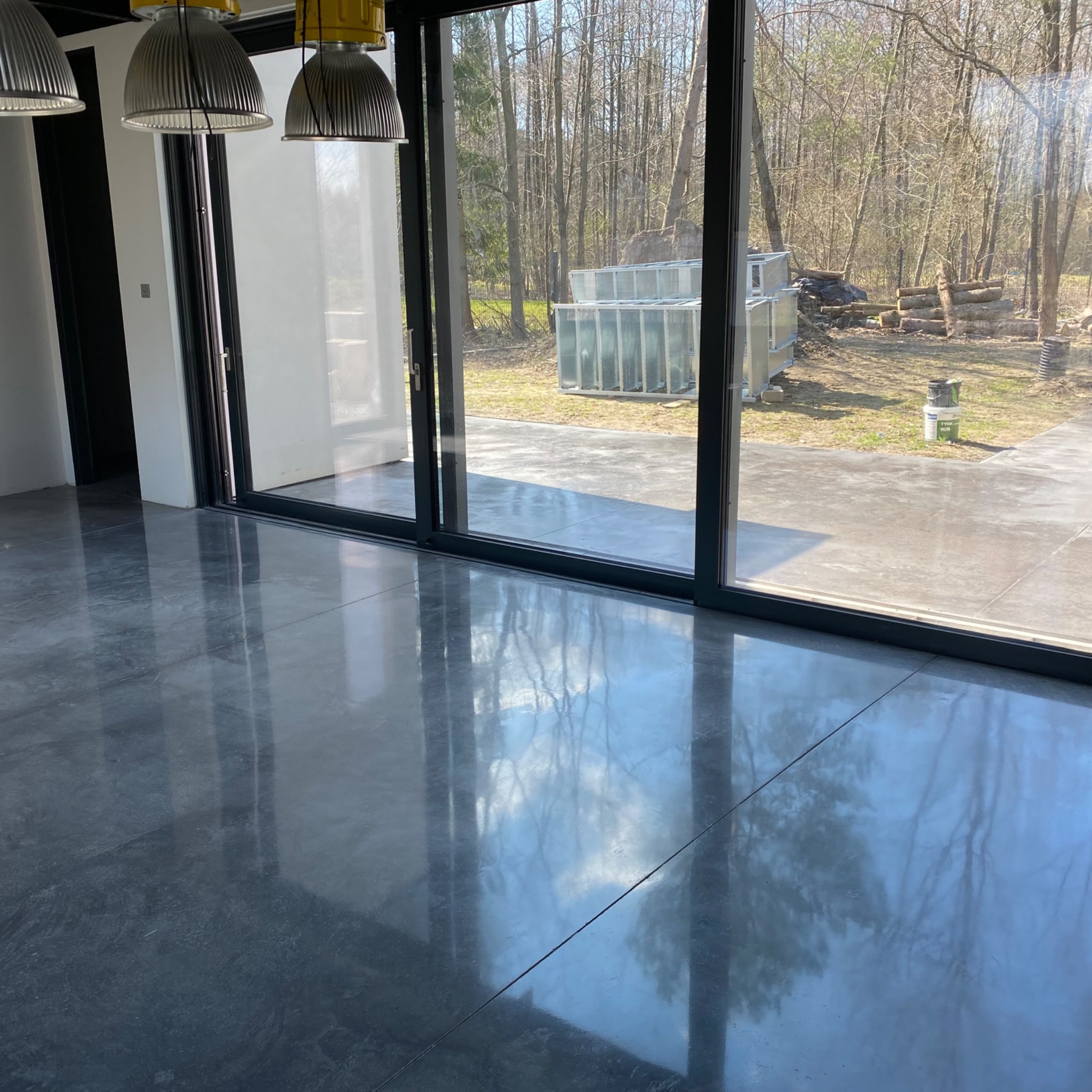 Błyszcząca, szara posadzka w nowoczesnym wnętrzu z dużym przesuwnym oknem, przez które widać drzewa i składowane materiały budowlane.