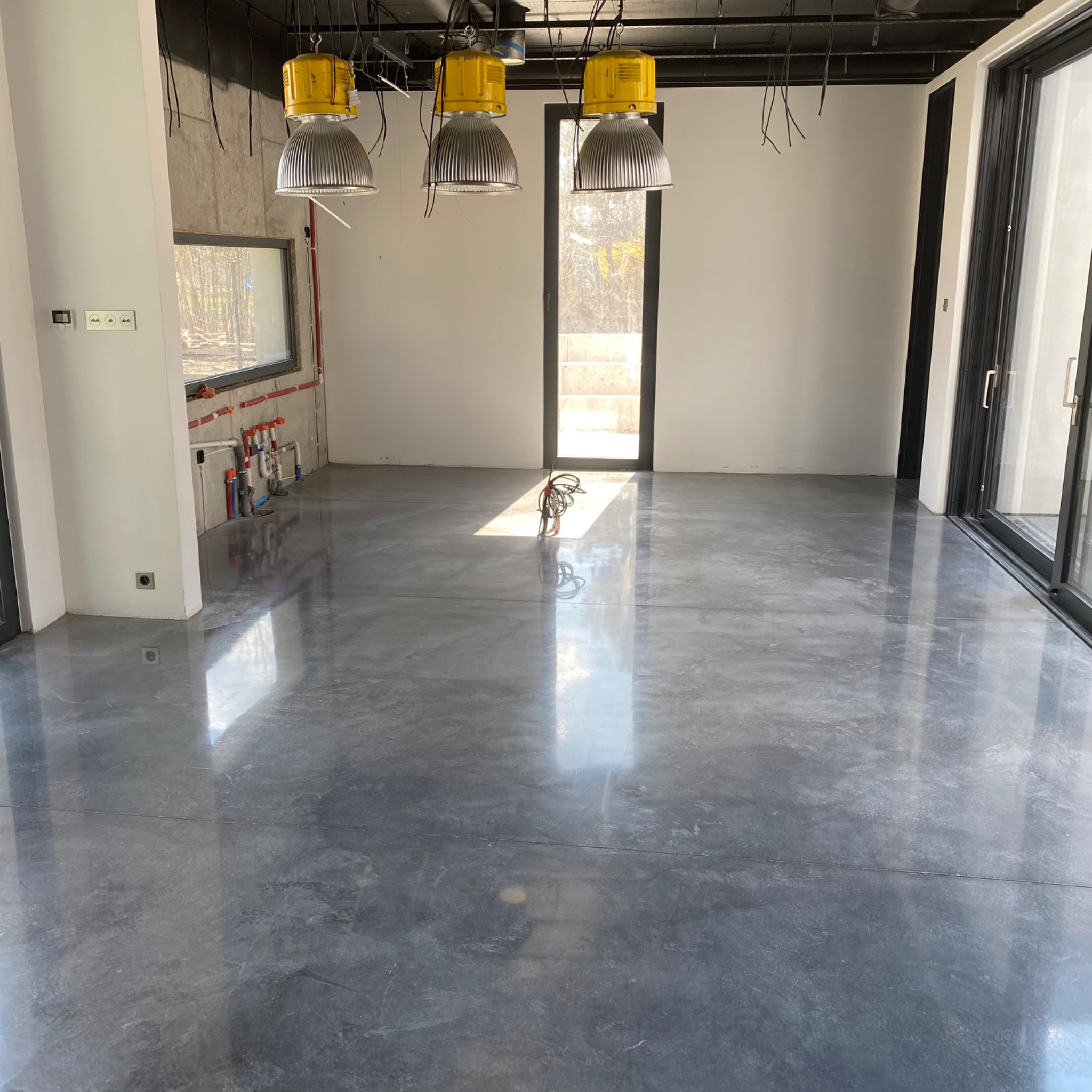 Błyszcząca, szara posadzka żywiczna w nowym domu w Justynowie, oświetlona przez trzy żółte lampy budowlane, z widocznymi instalacjami na ścianie i wiązką kabli na podłodze.