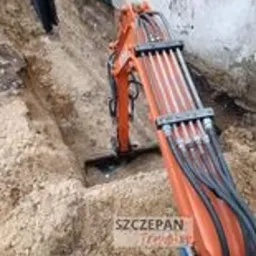 Pomarańczowa minikoparka w trakcie prac ziemnych, widoczny wykop w piaszczystej glebie, fragment budynku w tle.