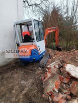 Mała koparka gąsienicowa Kubota w trakcie prac ziemnych, widoczna na tle gruzu ceglanego i drzew, z operatorem w kabinie.