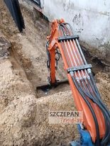 Pomarańczowa minikoparka w trakcie prac ziemnych, widoczny wykop w piaszczystej glebie, fragment budynku w tle.