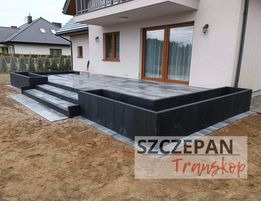 Nowoczesny taras z szarej kostki brukowej, zintegrowany z donicami z ciemnego drewna, prowadzący do jasnego domu z dużymi oknami tarasowymi. Widoczne schody z kostki brukowej i niezabudowany teren...