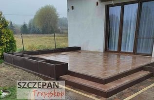 Brązowy taras wyłożony płytkami imitującymi drewno, z dwoma stopniami i podwyższonymi rabatami na rośliny, przylegający do jasnego budynku z dużymi oknami tarasowymi. Widoczne ogrodzenie posesji...