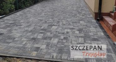 Ułożona szara kostka brukowa w geometryczny wzór, widoczny fragment budynku z rynną, obrzeża z odpływem liniowym, siatka ogrodzeniowa w tle.