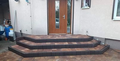 Trzystopniowe schody wejściowe do domu, wyłożone kostką brukową w odcieniach brązu i grafitu, z widocznym fragmentem elewacji i drzwiami wejściowymi z szybą.