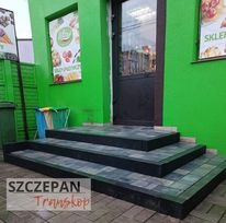 Trzy stopnie schodów wejściowych do sklepu wyłożone szarą kostką brukową z czarnym obrzeżem, zielona elewacja budynku w tle.