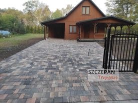 Ułożona kostka brukowa w odcieniach szarości i brązu prowadząca do drewnianego domu z podjazdem, widoczna brama wjazdowa z metalu, tło stanowią drzewa i krzewy.