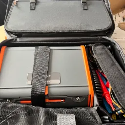 Otwarta torba elektryka z szarym miernikiem z pomarańczowymi elementami, czarnymi kablami i czerwono-niebieskimi sondami pomiarowymi w przegródkach.