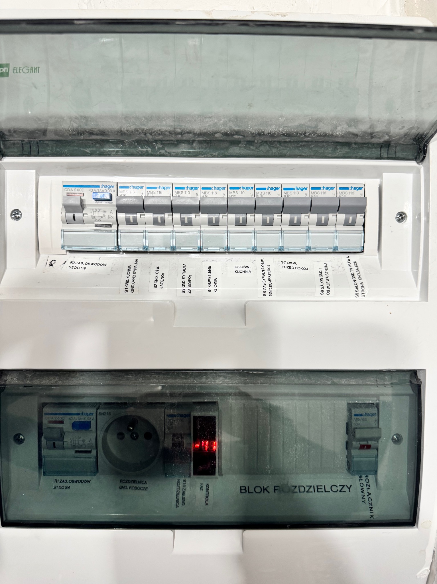 Otwarta skrzynka rozdzielcza Hager z bezpiecznikami i opisami obwodów (kuchnia, sypialnia, salon). Widoczny rozłącznik i gniazdo. Blok rozdzielczy wewnątrz budynku.