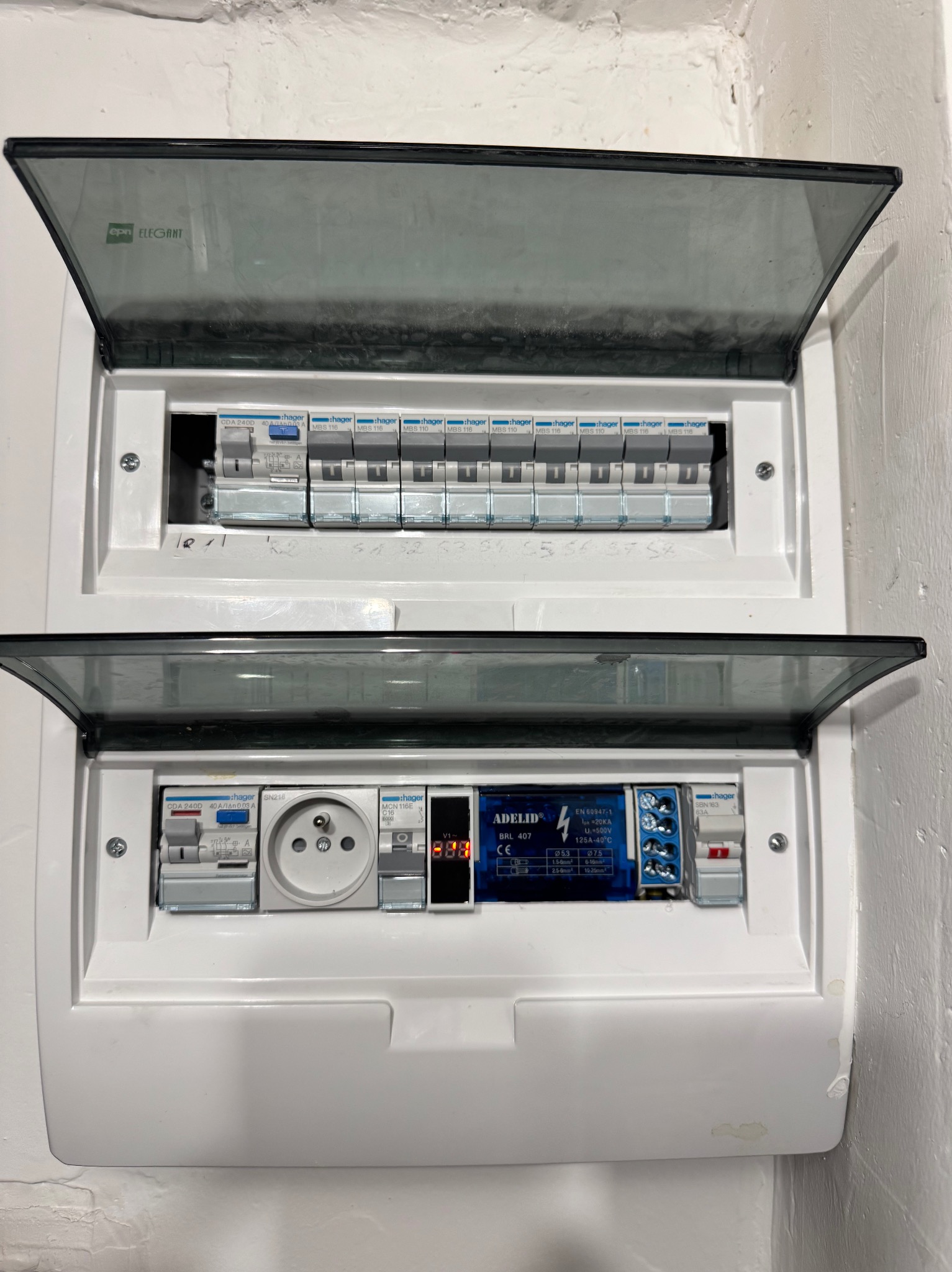 Rozdzielnica elektryczna Hager z otwartymi klapami, widoczne zabezpieczenia, gniazdo i moduł Adelid BRL 407. Biała obudowa na tle ściany.