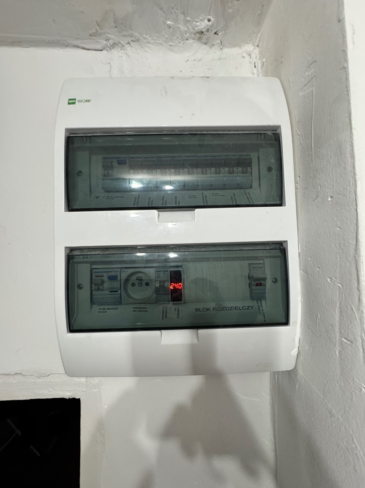 Biała skrzynka rozdzielcza z widocznymi bezpiecznikami, gniazdem i wyświetlaczem napięcia 240V, zamontowana na białej, nierównej ścianie. Widoczny napis 'Blok Rozdzielczy'.