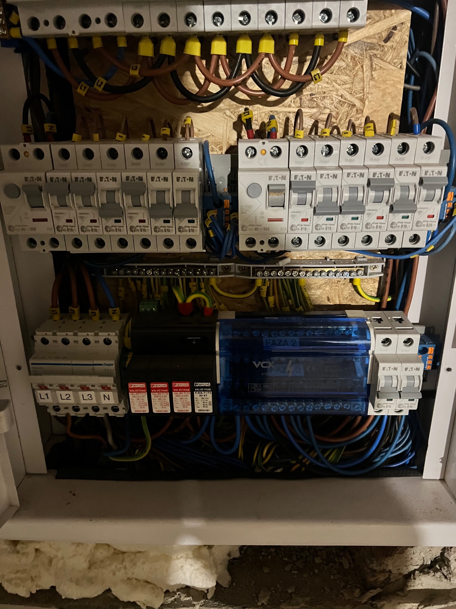 Rozdzielnica elektryczna z zabezpieczeniami Eaton, widoczne bezpieczniki, przewody oznaczone kolorami i numerami, zamontowana na płycie OSB wewnątrz obudowy.