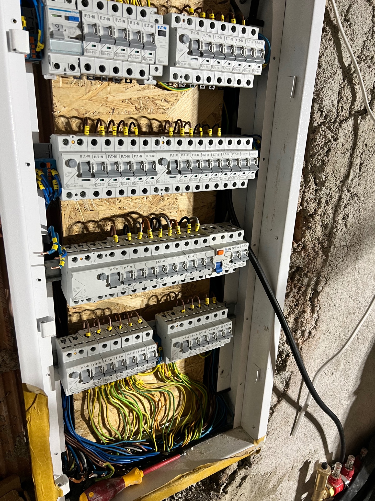 Rozdzielnica elektryczna z zamontowanymi bezpiecznikami EATON, widoczne przewody w różnych kolorach. Instalacja w trakcie prac, surowa ściana w tle.