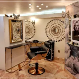 Eleganckie wnętrze z fotelami fryzjerskimi, toaletkami z marmurowymi blatami i dekoracyjnymi lustrami w stylu glamour. Na ścianach ozdobne lustra w kształcie słońca i portret Marilyn Monroe.