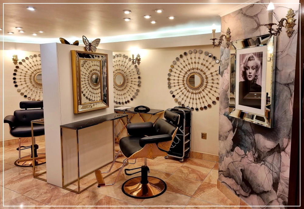 Eleganckie wnętrze z fotelami fryzjerskimi, toaletkami z marmurowymi blatami i dekoracyjnymi lustrami w stylu glamour. Na ścianach ozdobne lustra w kształcie słońca i portret Marilyn Monroe.