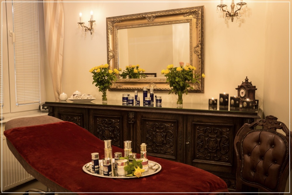 Ekskluzywny gabinet z leżanką pokrytą czerwonym aksamitem, srebrną tacą z kosmetykami, ozdobnym kredensem z lustrem w złotej ramie, bukietami żółtych róż i świecami.
