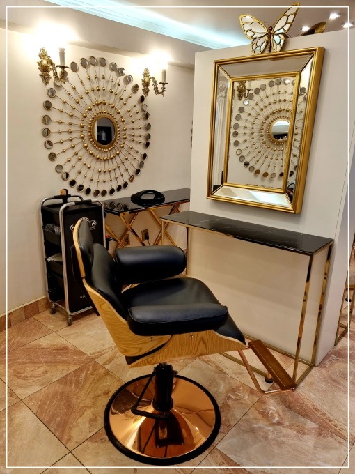 Nowoczesne wnętrze: fotel fryzjerski z czarną tapicerką i drewnianym oparciem, złote lustro z motylem, dekoracyjne lustro na ścianie i złote konsole z czarnym blatem.