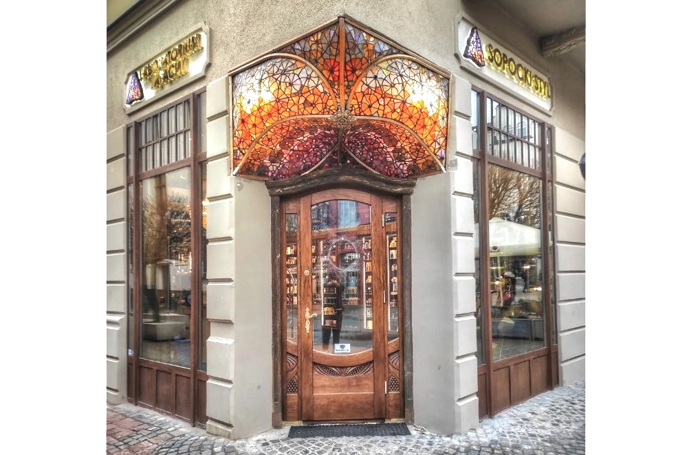 Fasada budynku z drewnianymi oknami i drzwiami, nad wejściem witraż w odcieniach czerwieni i pomarańczy, szyldy reklamowe po bokach.