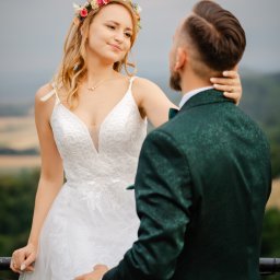 RADOSŁAW WASILEWSKI FOTOFIZYK - Para młoda: panna młoda w białej sukni z wiankiem spogląda na pana młodego w zielonym smokingu, na tle rozmytego krajobrazu. Romantyczna sesja w plenerze.