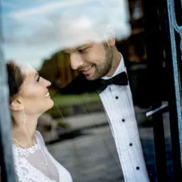 Panna młoda w koronkowej sukni i pan młody w smokingu z muchą, patrzący na siebie przez ozdobne, metalowe okno z refleksami.