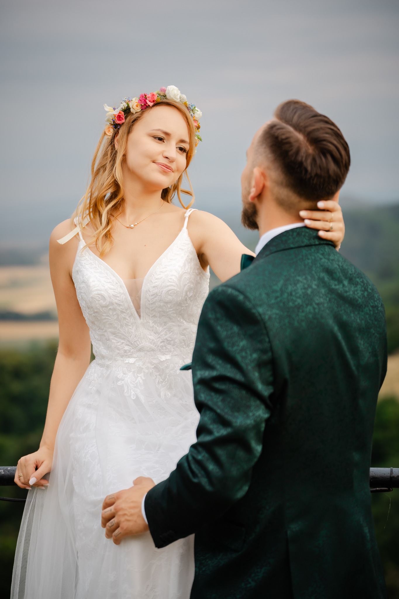 Para młoda: panna młoda w białej sukni z wiankiem spogląda na pana młodego w zielonym smokingu, na tle rozmytego krajobrazu. Romantyczna sesja w plenerze.