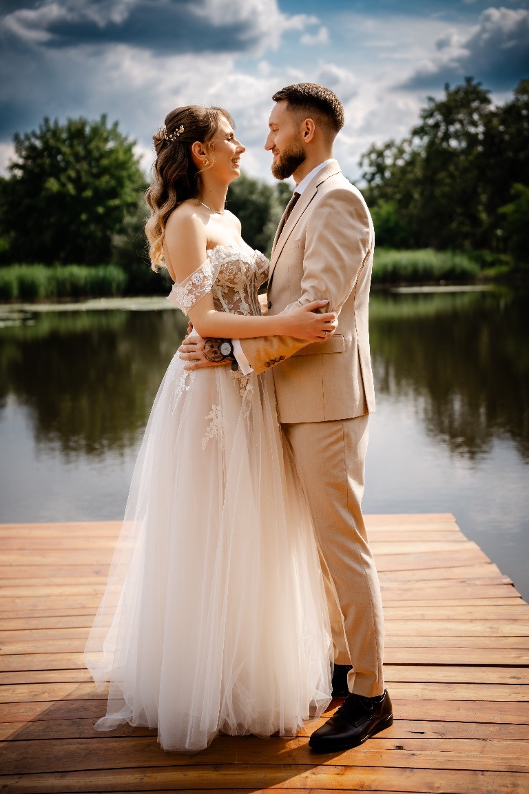 Para młoda w beżowych strojach, obejmująca się na drewnianym pomoście nad jeziorem, w tle zieleń drzew i błękitne niebo z chmurami. Romantyczna sesja plenerowa.