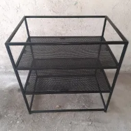 Metalowa konstrukcja regału z trzema ażurowymi półkami z siatki, spawana z czarnych profili, na surowym betonowym podłożu.