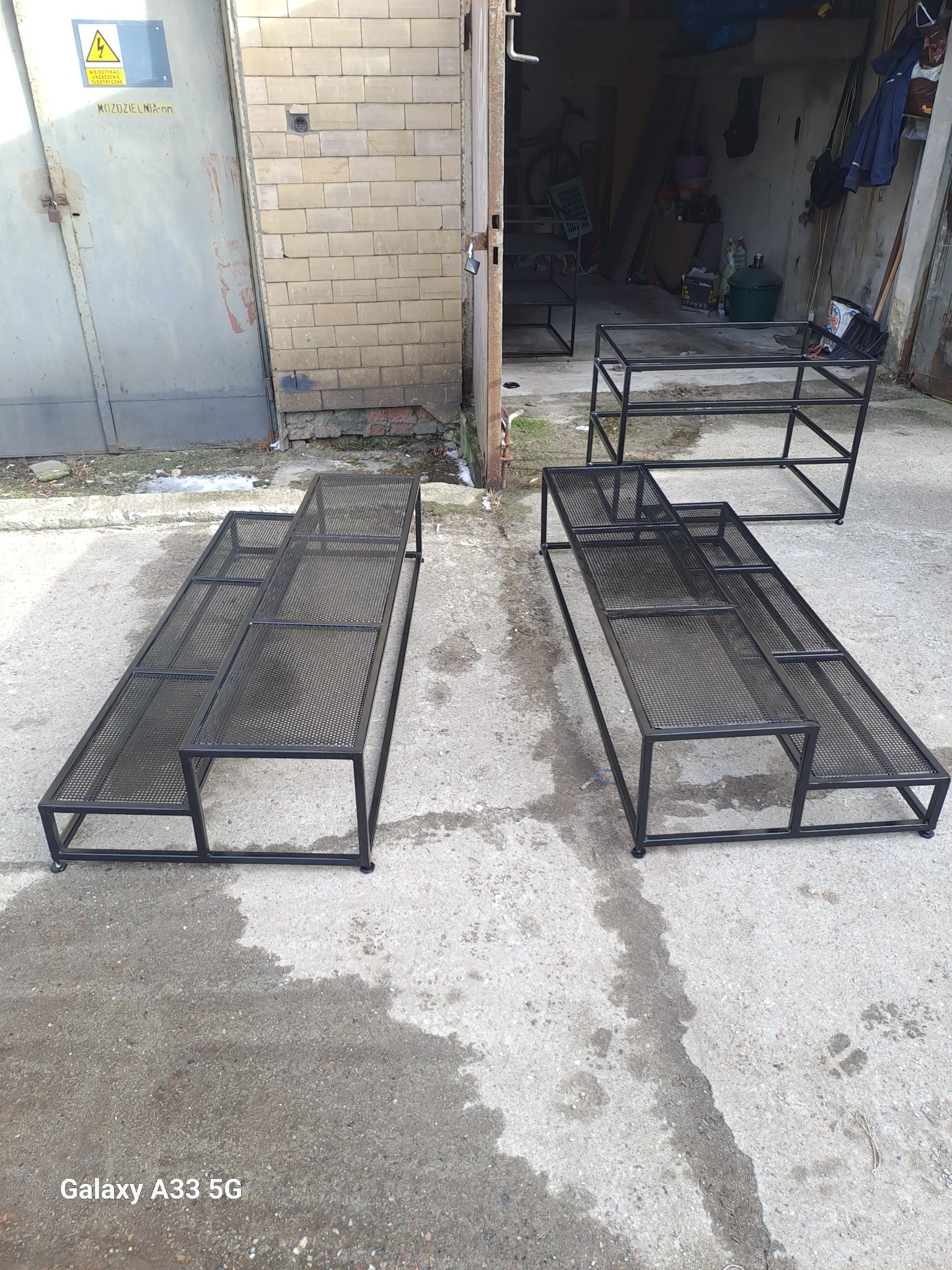 Metalowe konstrukcje na zewnątrz warsztatu, dwie długie, schodkowe platformy z siatki i mniejsza, prostokątna, prawdopodobnie przygotowane do malowania proszkowego.