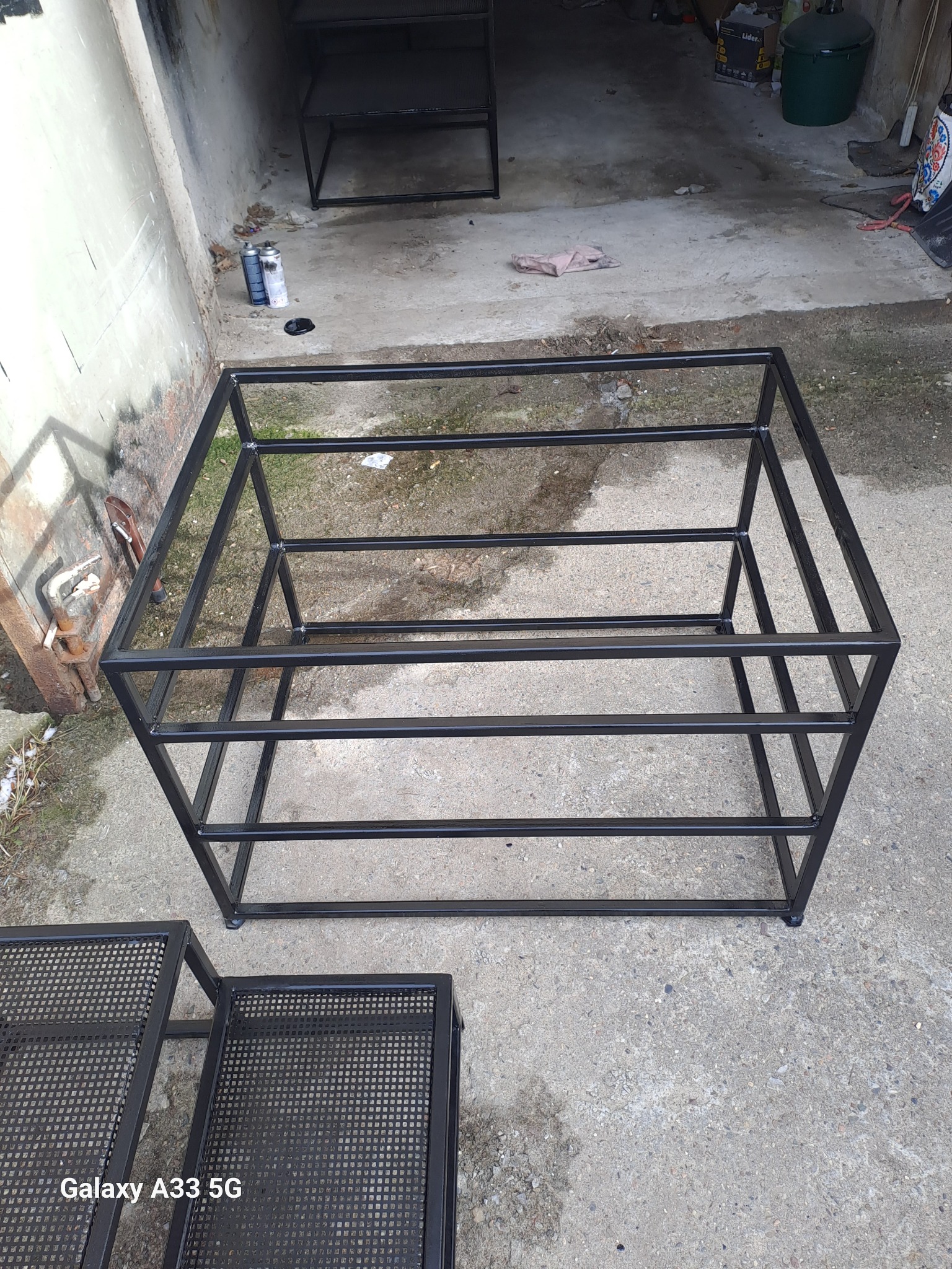 Surowa, spawana konstrukcja metalowa w kształcie prostopadłościanu z poziomymi wzmocnieniami, czarna, widoczne ślady spawania, w tle puszki farby i inne metalowe elementy, wykonana na zewnątrz.