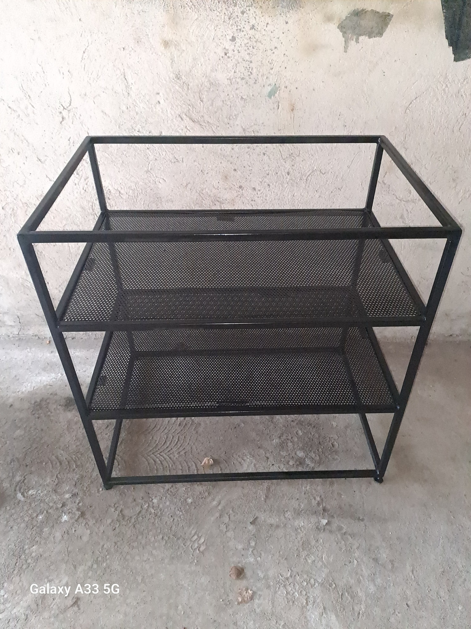 Metalowa konstrukcja regału z trzema ażurowymi półkami z siatki, spawana z czarnych profili, na surowym betonowym podłożu.