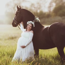 Żaneta Tarnowska Artistry - Kobieta w białej sukni ciążowej, z wiankiem na głowie, opiera się o czarnego konia z kwiatami na grzbiecie, na łące w ciepłym świetle.