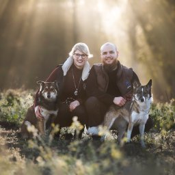 Żaneta Tarnowska Artistry - Uśmiechnięta para w kurtkach pozuje z dwoma psami rasy husky na łące w promieniach porannego słońca.