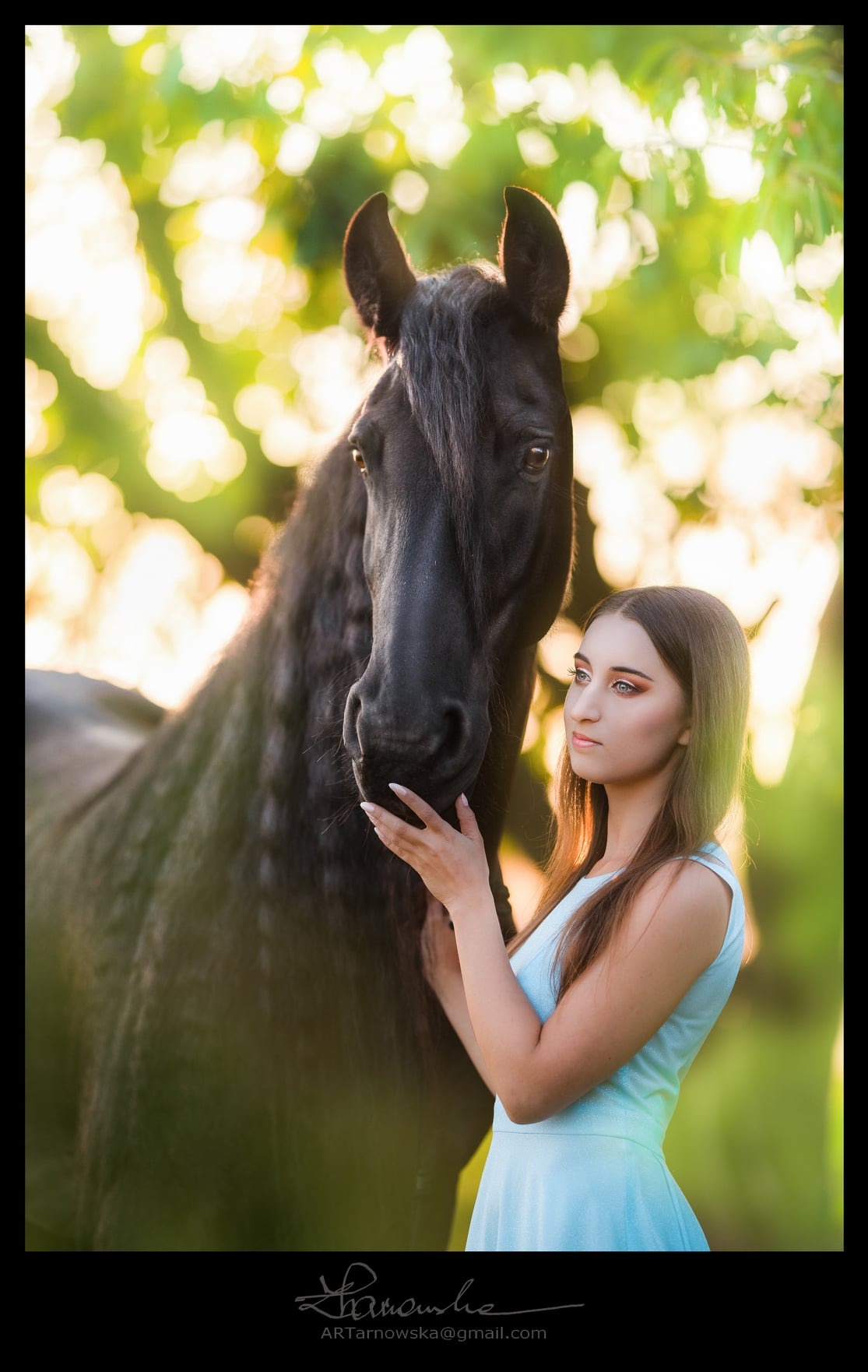 Portret młodej kobiety w błękitnej sukience, delikatnie dotykającej szyi czarnego konia na tle rozmytej zieleni i złotego światła.