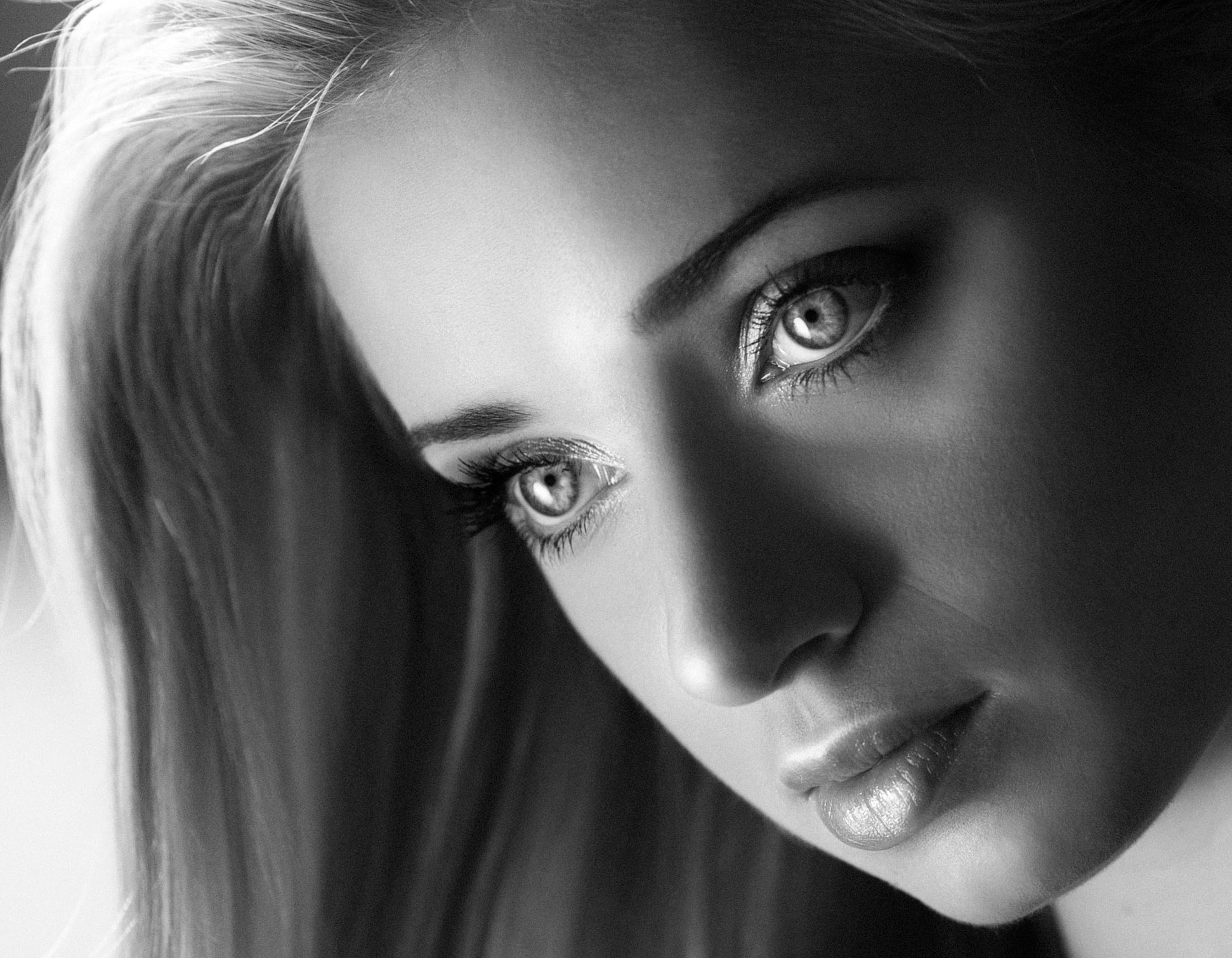 Czarno-białe zbliżenie na twarz młodej kobiety z długimi włosami, skupiające się na jej oczach i ustach, z delikatnym makijażem i subtelnym oświetleniem.