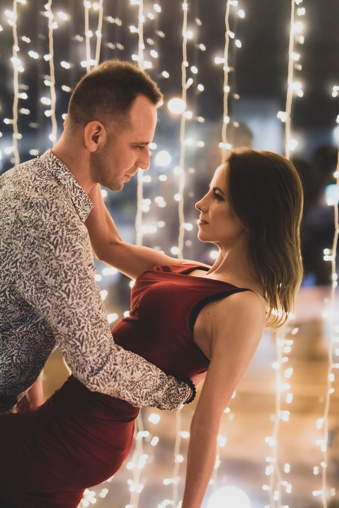 Para w objęciach, kobieta w czerwonej sukience odchyla się do tyłu, mężczyzna w koszuli we wzory patrzy na nią, tło rozmyte z dekoracyjnych światełek.
