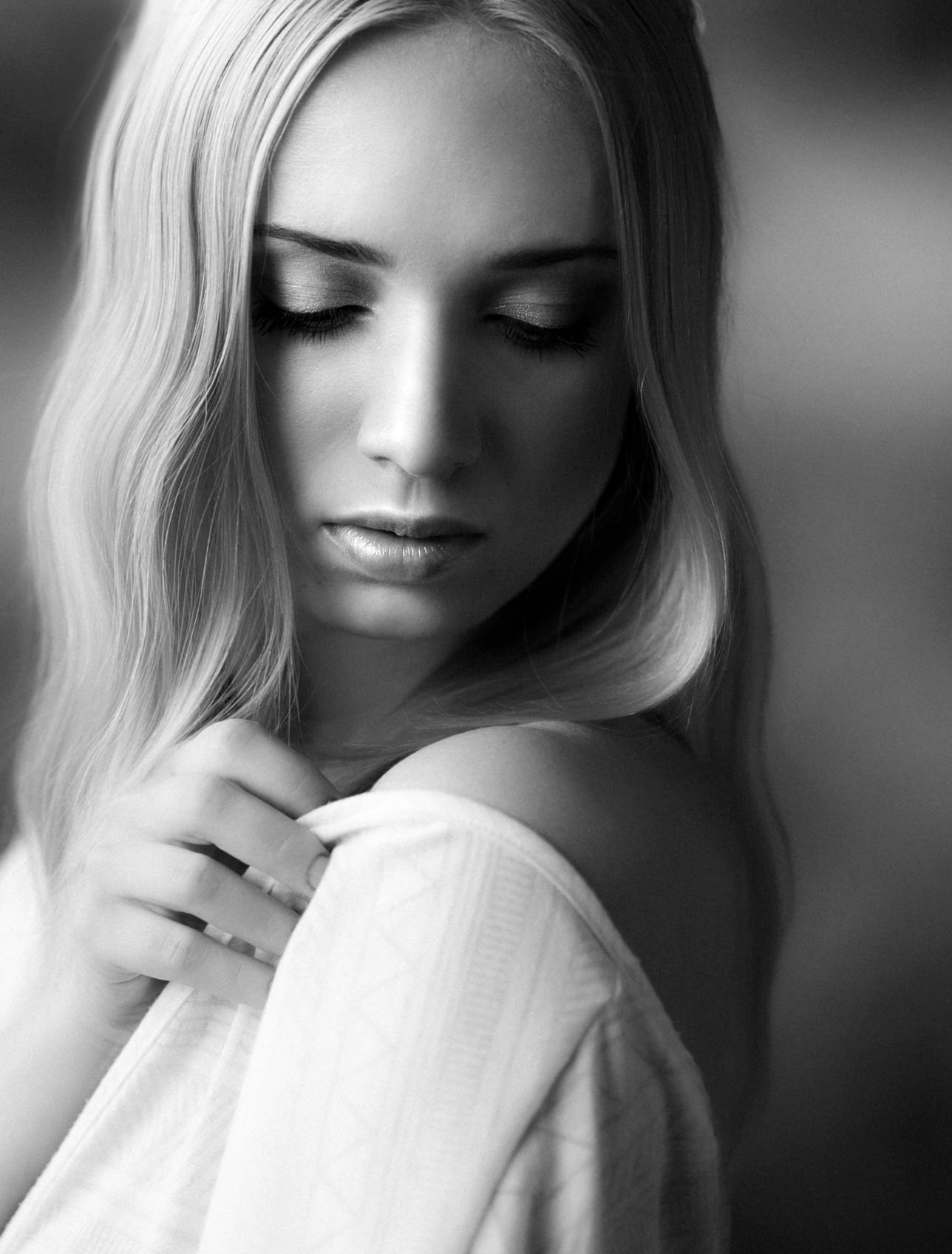 Portret w czerni i bieli młodej kobiety z długimi blond włosami, delikatny makijaż, spuszczony wzrok, dłoń na ramieniu, subtelne światło.